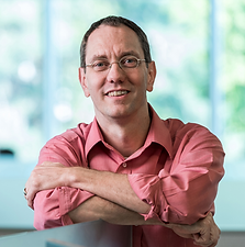 A man with glasses and short hair, wearing a pink shirt, smiling with his arms crossed, sitting in a bright room with large windows and greenery outside.