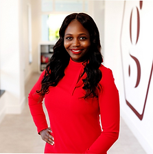 A woman with dark hair and a bright smile wearing a red long-sleeve top standing in a hallway with artwork on the wall.
