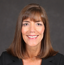 A smiling woman with shoulder-length brown hair and bangs, wearing a black blazer against a gray background.