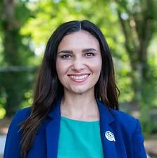 A woman with long dark hair smiling outdoors, wearing a blue blazer and teal top.