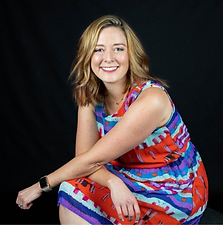 A young woman with shoulder-length blonde hair and a bright smile, wearing a colorful patterned dress, sitting against a black background.