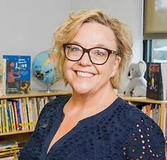A smiling woman with glasses and short blonde hair in front of a bookshelf with children's books and stuffed animals.