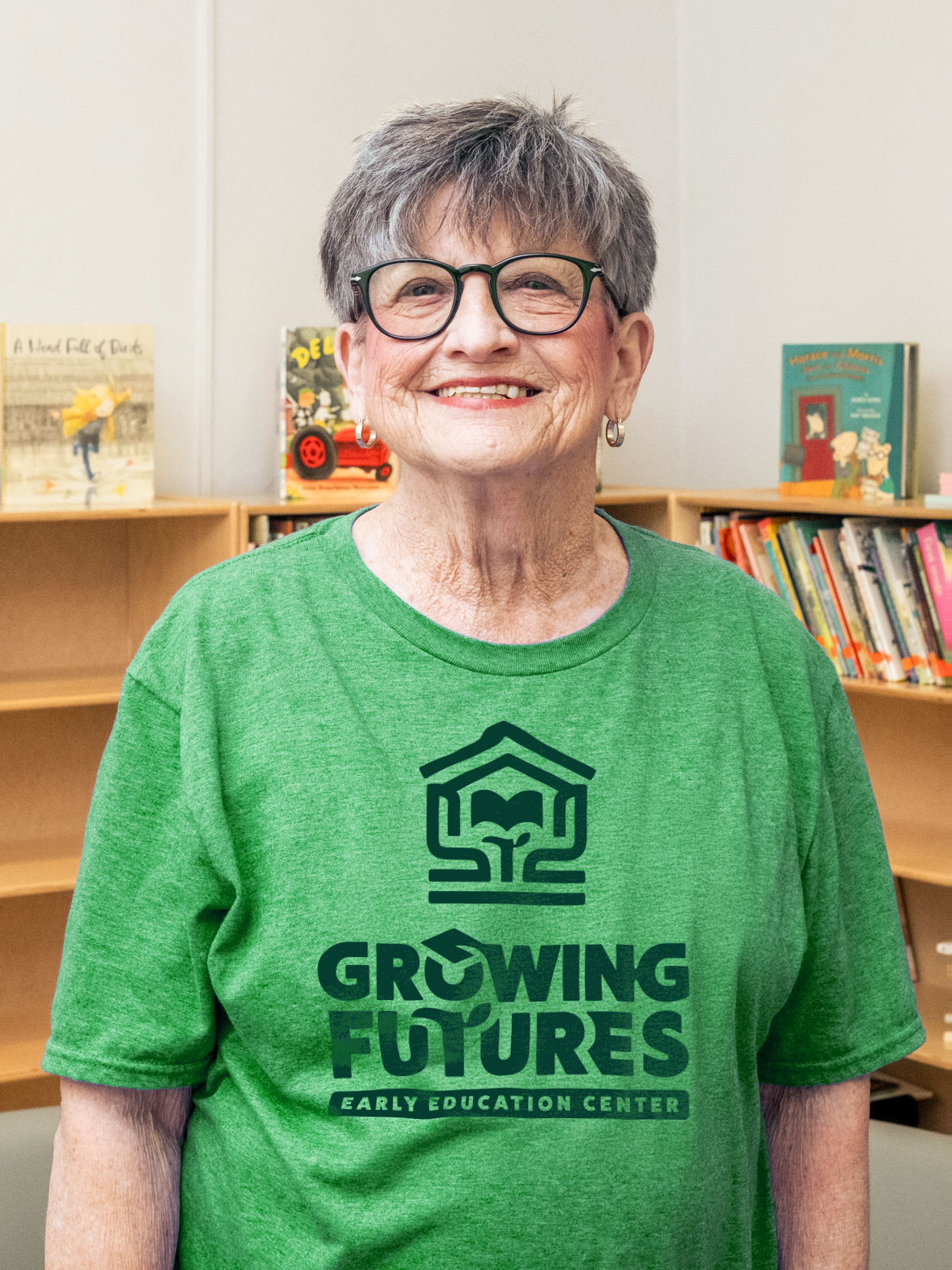An older woman with short gray hair, glasses, and earrings, smiling while wearing a green t-shirt that says 'Growing Futures Early Education Center,' standing in front of bookshelves filled with children's books.