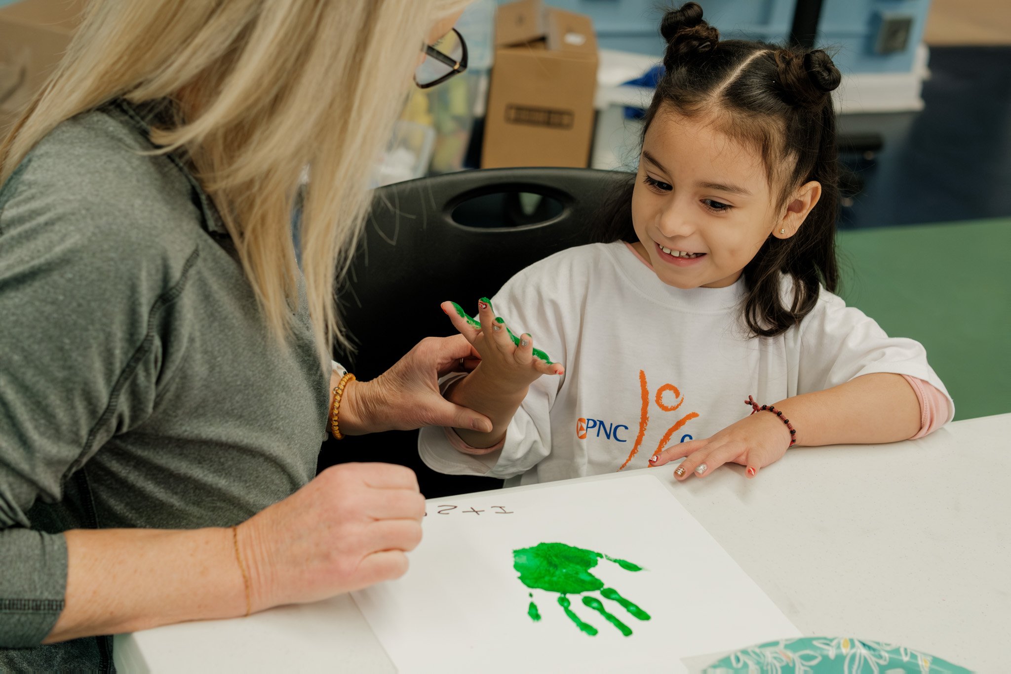 A young girl with dark hair styled in two buns smiling while showing her painted hand to an adult woman with blonde hair, both sitting at a table with a white paper displaying a green handprint and some math in a classroom or art room.