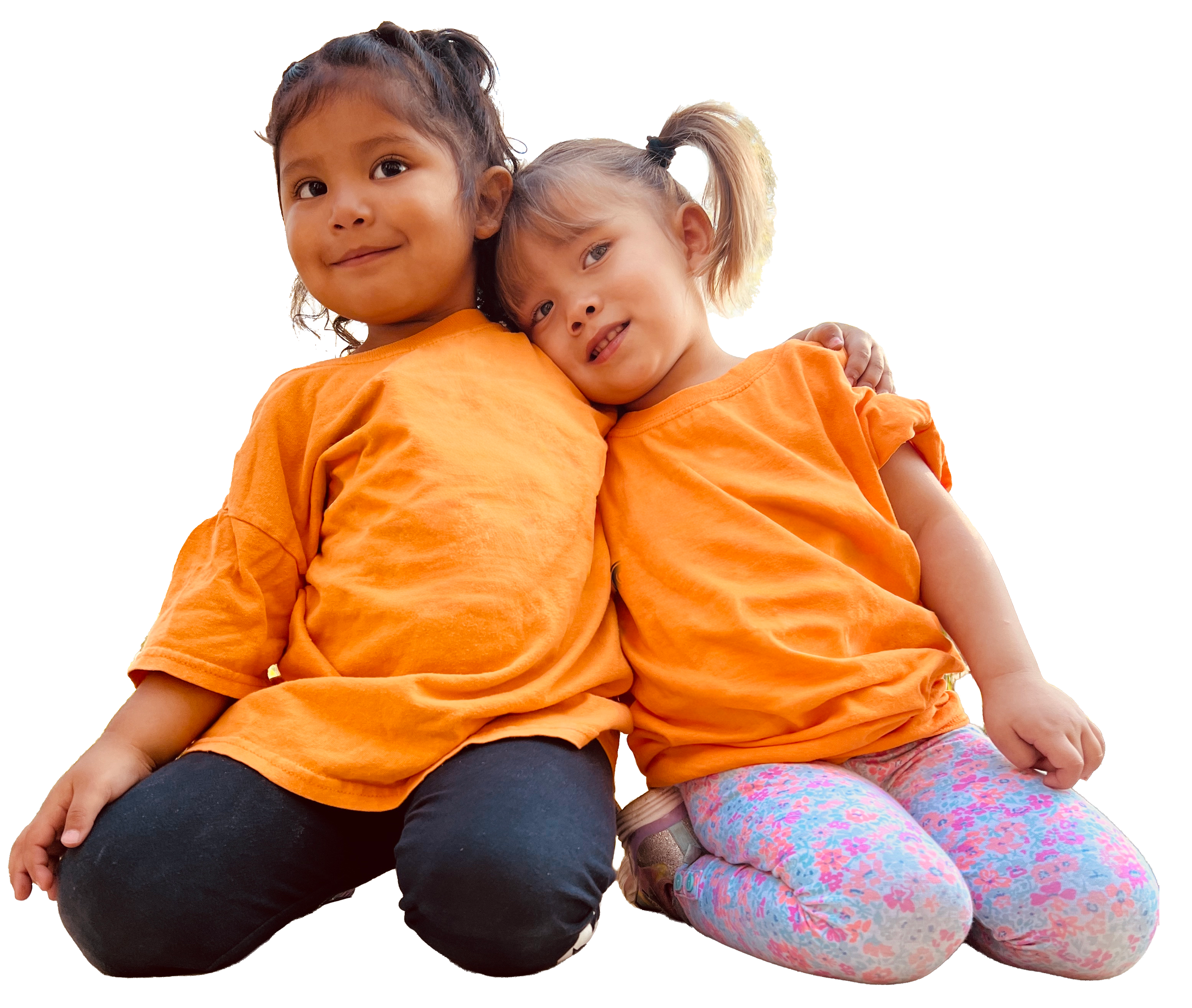 Two young girls sitting close together, both wearing orange shirts. The girl on the left has darker skin and dark hair tied back, smiling slightly. The girl on the right has lighter skin and blonde hair in pigtails, leaning her head against the other girl, smiling with her teeth visible.
