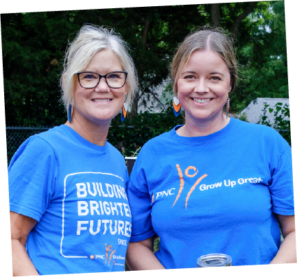 Two women smiling outdoors, wearing matching blue T-shirts with logos and slogans, standing in front of green trees.