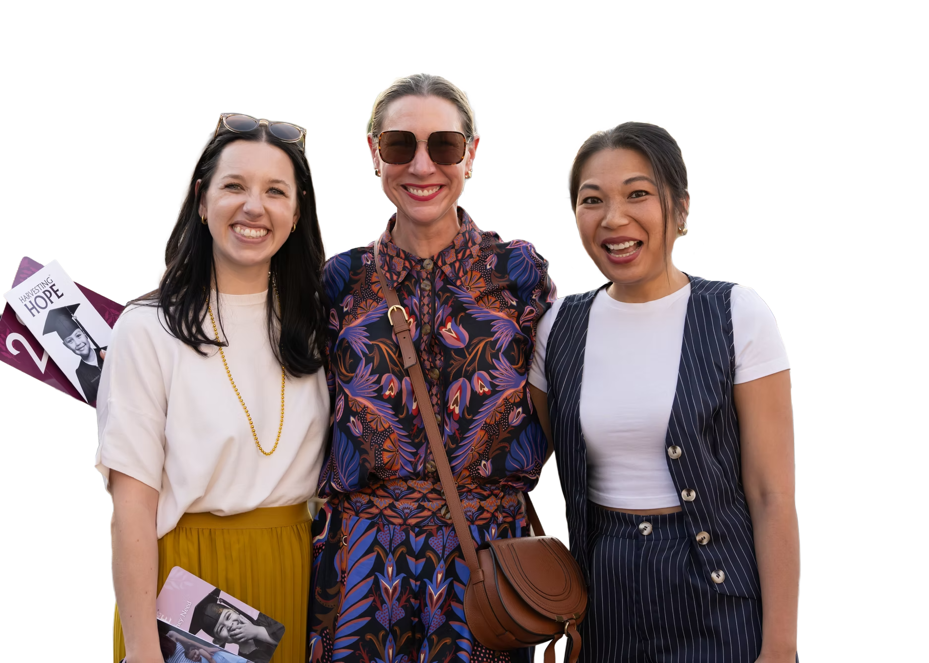 Three women smiling and standing close together outdoors, one holding a graduation program. The woman on the left has long dark hair, wearing a white blouse and yellow skirt. The woman in the middle has blonde hair, sunglasses, and is wearing a multicolored printed dress. The woman on the right has dark hair, wearing a white shirt with a pinstriped vest and matching skirt.
