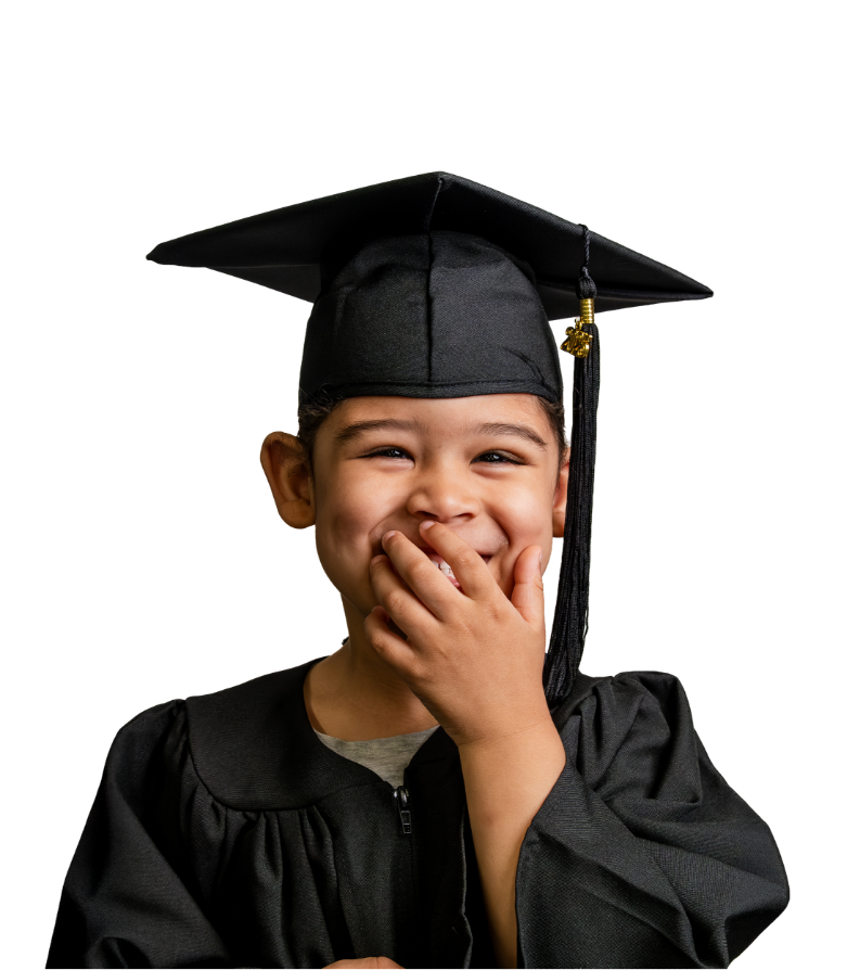 A young boy in a black graduation cap and gown, smiling and covering his mouth with his hand.