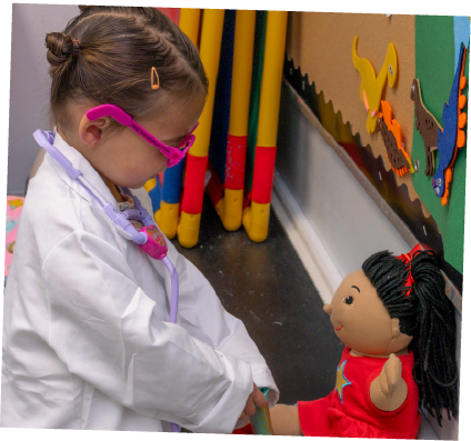 A young girl dressed as a doctor playing with a doll in a classroom with educational wall decorations.
