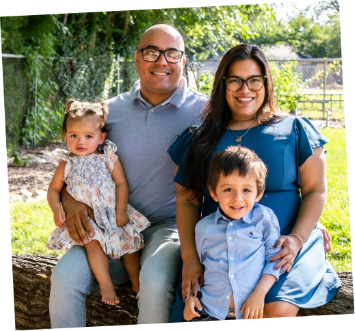 Family of four sitting outdoors on a log, smiling at the camera. The father, wearing glasses and a gray polo shirt, sits next to a woman in a blue dress. Their young daughter, in a floral dress, sits on the father's lap. Their young son, in a light blue shirt, sits on the woman's lap. Green trees and a chain-link fence are in the background.