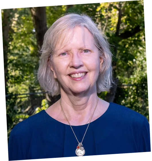 A smiling woman with blonde hair outdoors, wearing a blue top and a silver necklace, surrounded by green trees and sunlight.