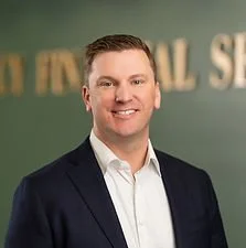 A smiling man in a dark suit and white shirt standing in front of a green background with text.