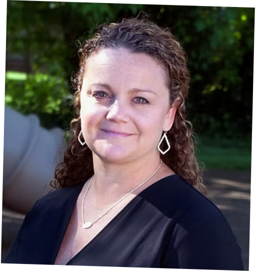 A woman with curly brown hair standing outdoors with greenery in the background, wearing a black top and jewelry.