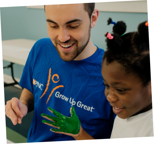 A young man with a beard and a girl with braided hair are smiling. The man is wearing a blue shirt with the PNC logo and the phrase "Grow Up Great." The girl has green paint on her hand, which she is showing to the man.