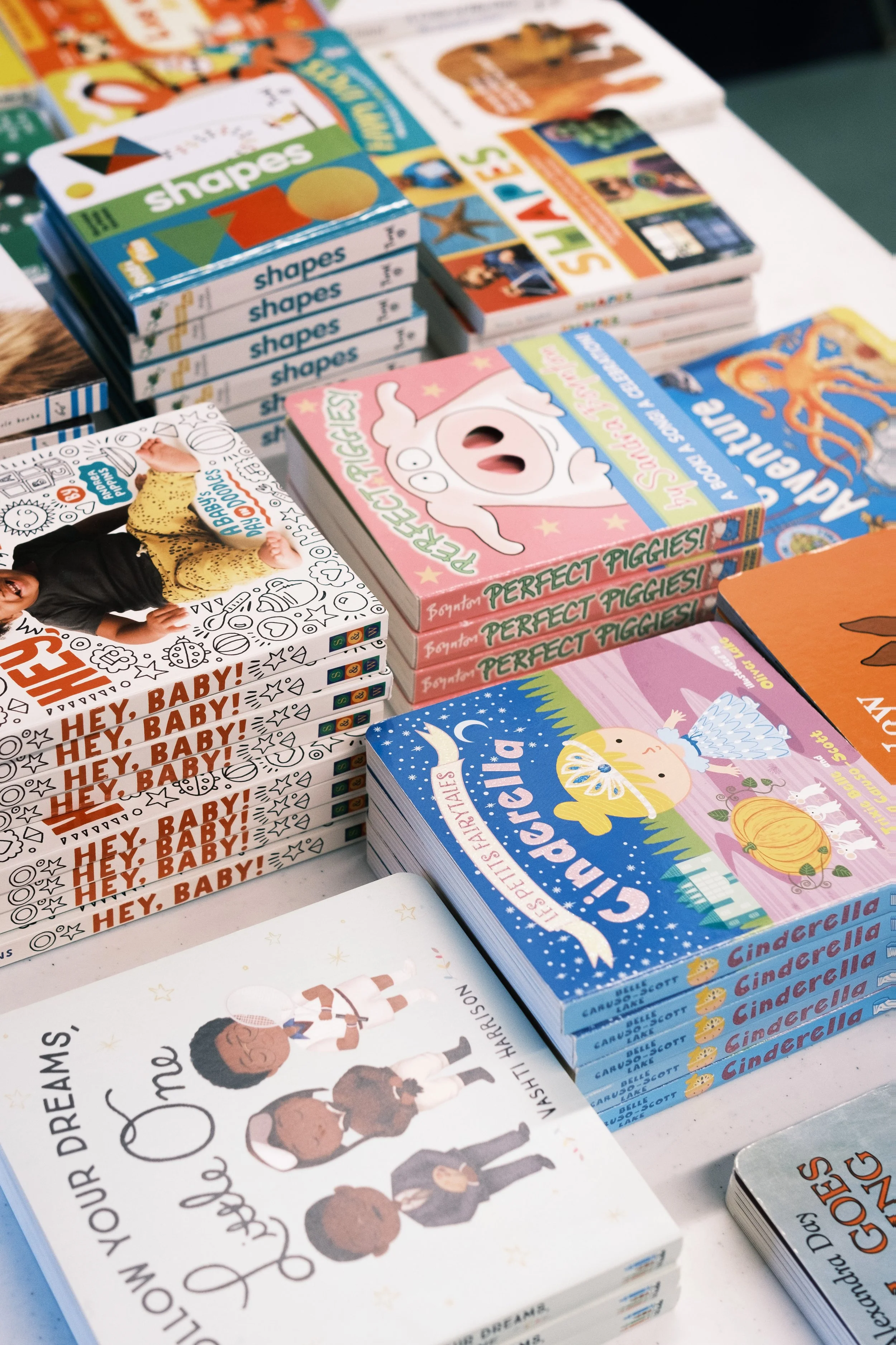 Stacked children's books and activity books on a table, including titles like 'Cinderella', 'Perfect PIGGIES!', 'Hey, Baby!', and educational books about shapes and colors.