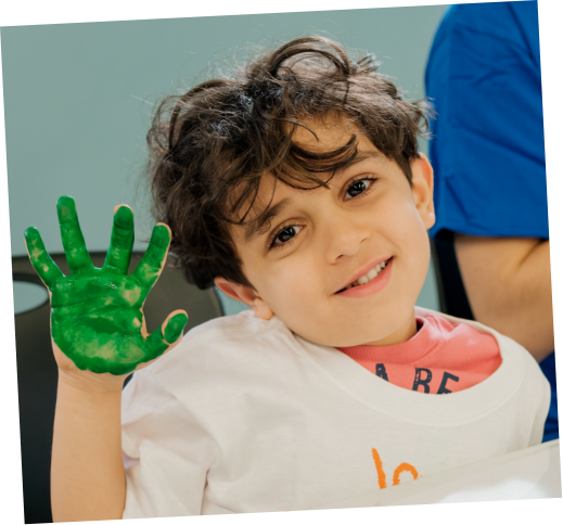 Young boy with brown curly hair smiling, raising his hand with green painted fingers.