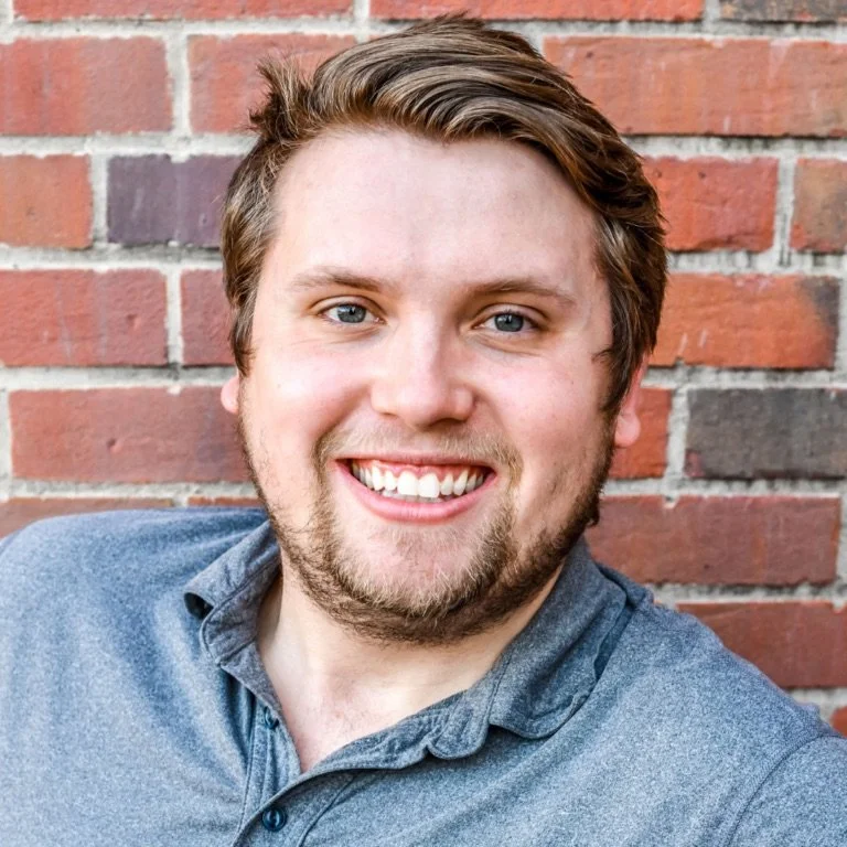 A smiling young man with light brown hair, a beard, and blue eyes, wearing a blue denim shirt, posing outdoors in front of a brick wall.