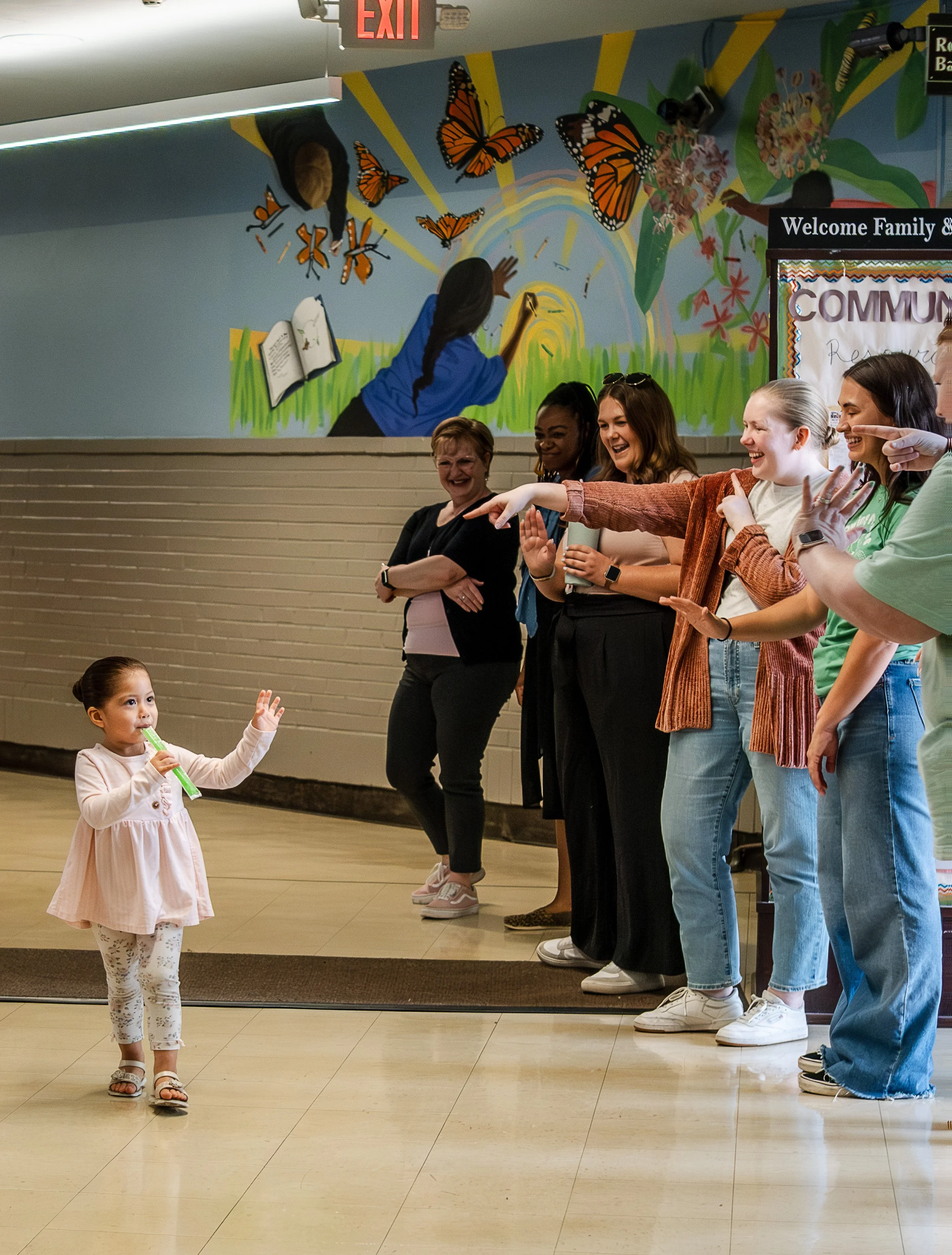 A young girl playing a recorder in front of a group of smiling women in a hallway with a colorful mural of a girl drawing butterflies on the wall.