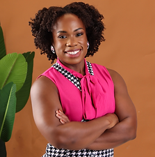 A smiling woman with curly hair wearing a pink sleeveless top with black and white checkered collar, standing with arms crossed next to a green plant against a beige background.