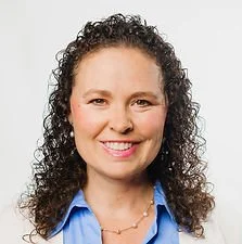 A woman with curly dark hair smiling, wearing a blue collared shirt and a necklace.