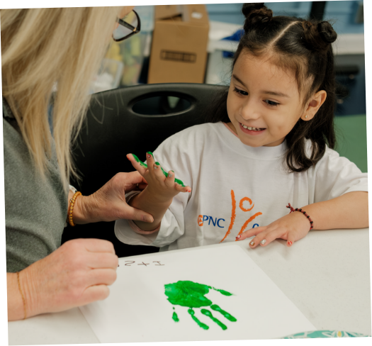 A young girl receiving a handprint painted with green paint from an adult at a table in a classroom or art room.