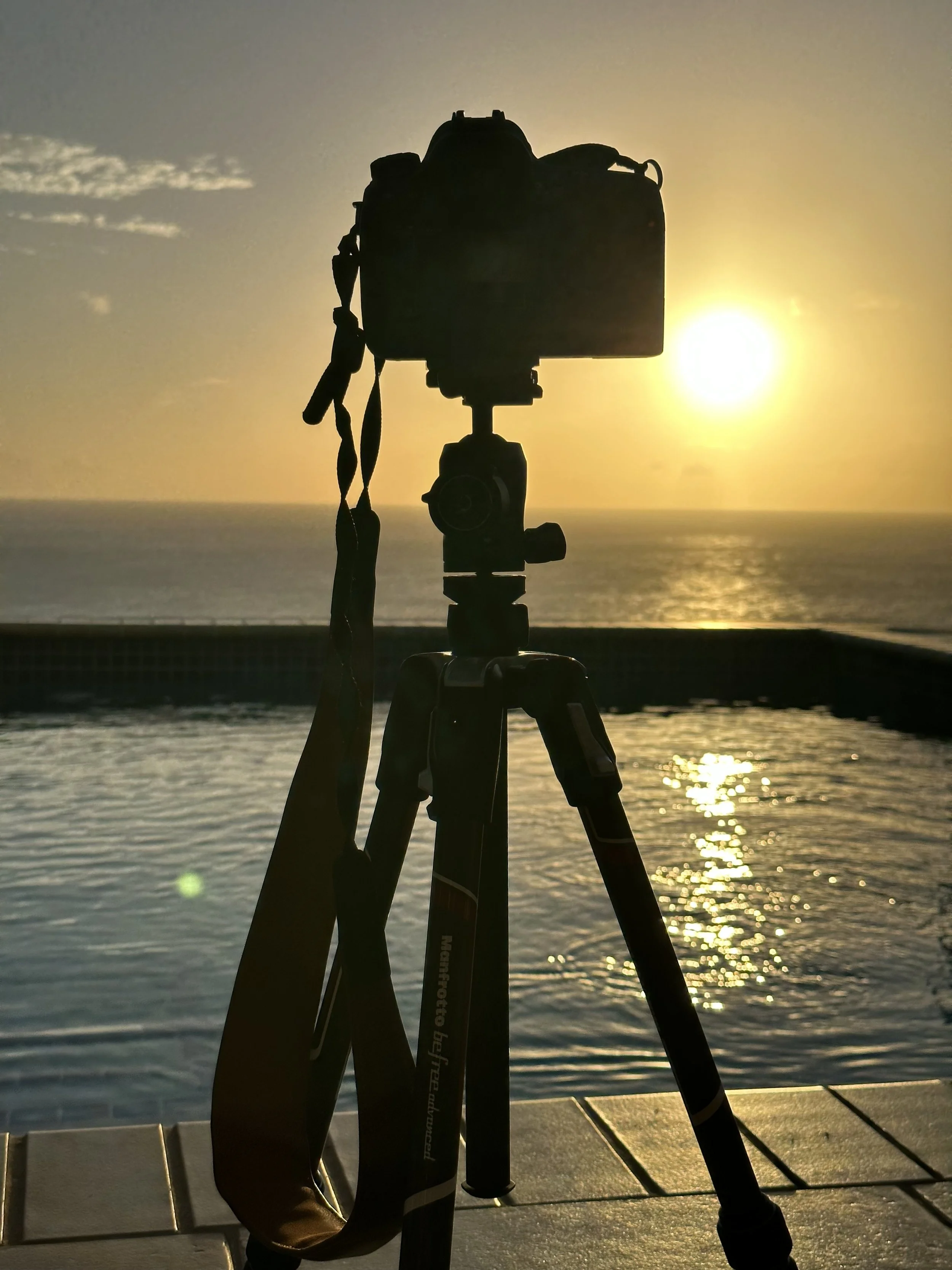 Silhouette of a camera on a tripod in front of a sunset over a body of water.