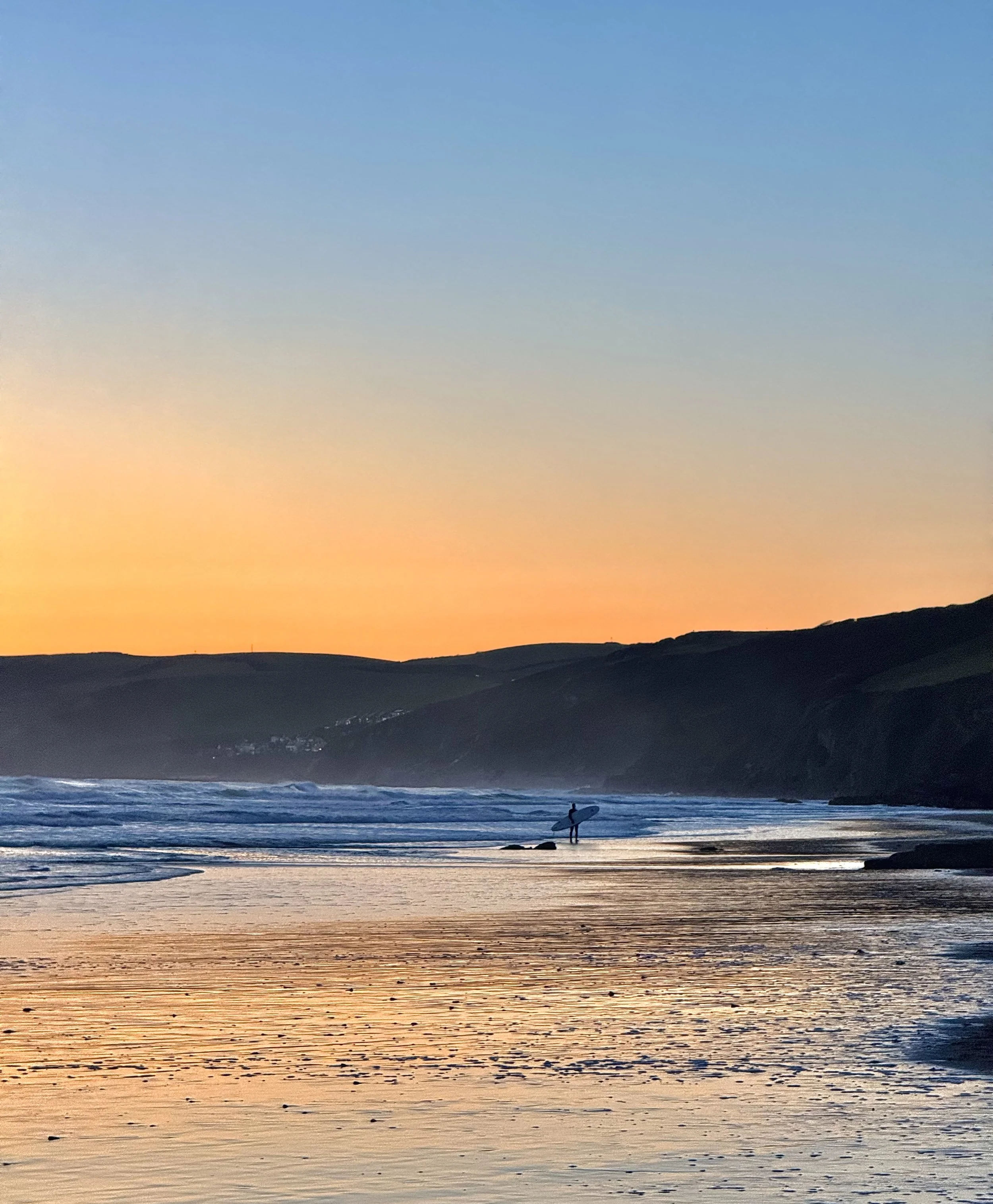 Silhouette of a person holding a surfboard on a beach during sunset with colorful sky and distant hills