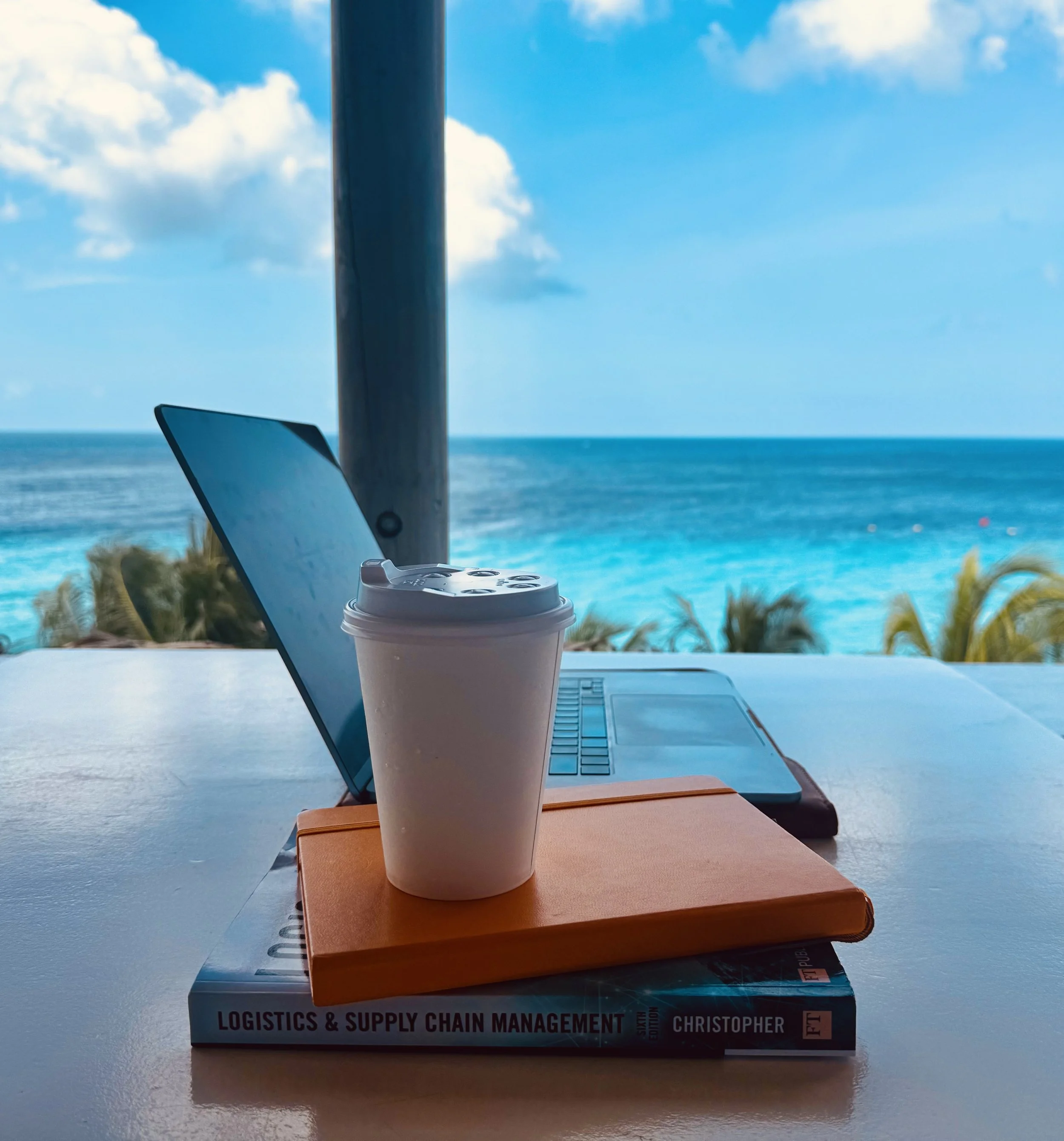 A book titled "Logistics & Supply Chain Management" by Christopher lying on a table with a laptop, a paper coffee cup, and a closed laptop in front of a window showing a view of the ocean and blue sky with clouds.