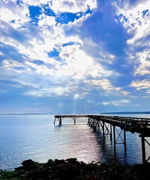 A pier extends into a calm body of water under a partly cloudy sky with sun rays shining through the clouds.