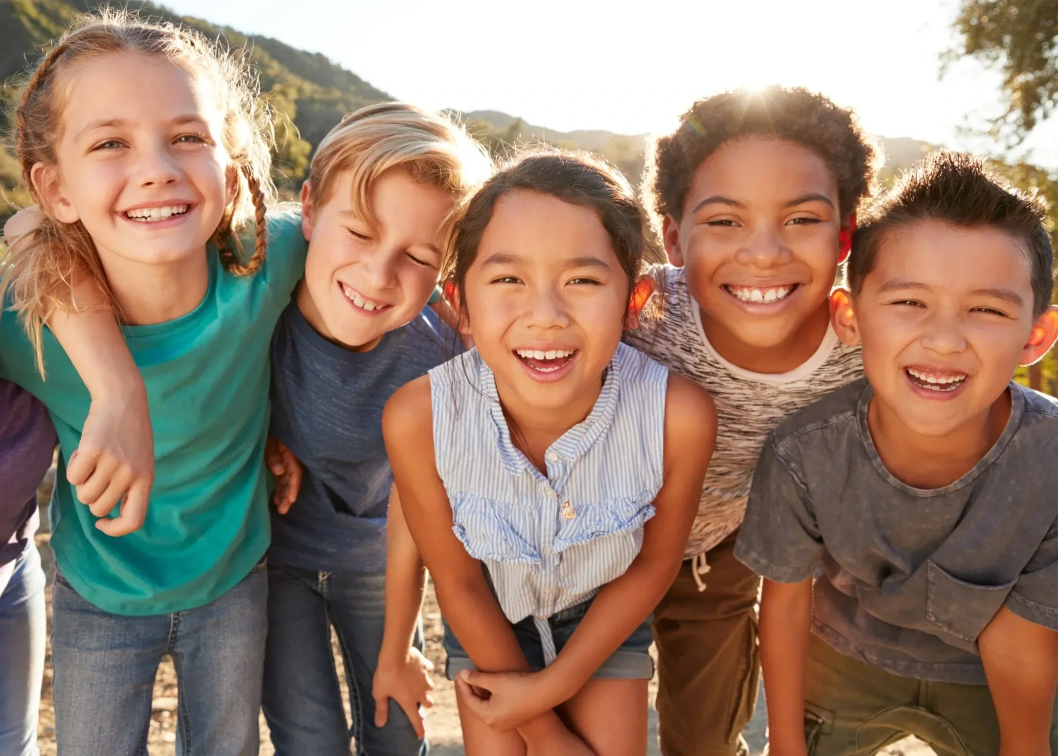 A group of five kids smiling and leaning into each other outdoors