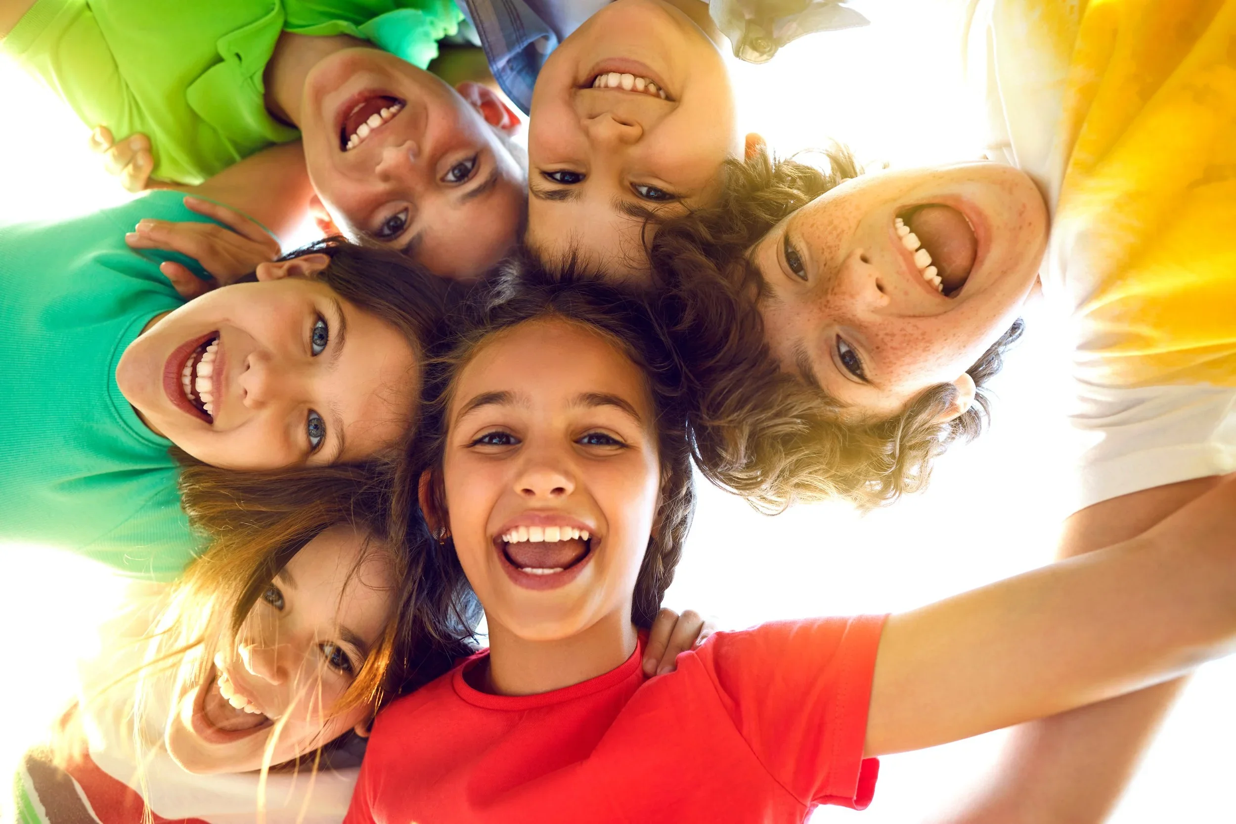 A close-up of smiling children in a circle, looking up at the camera — bright, joyful, peer connection