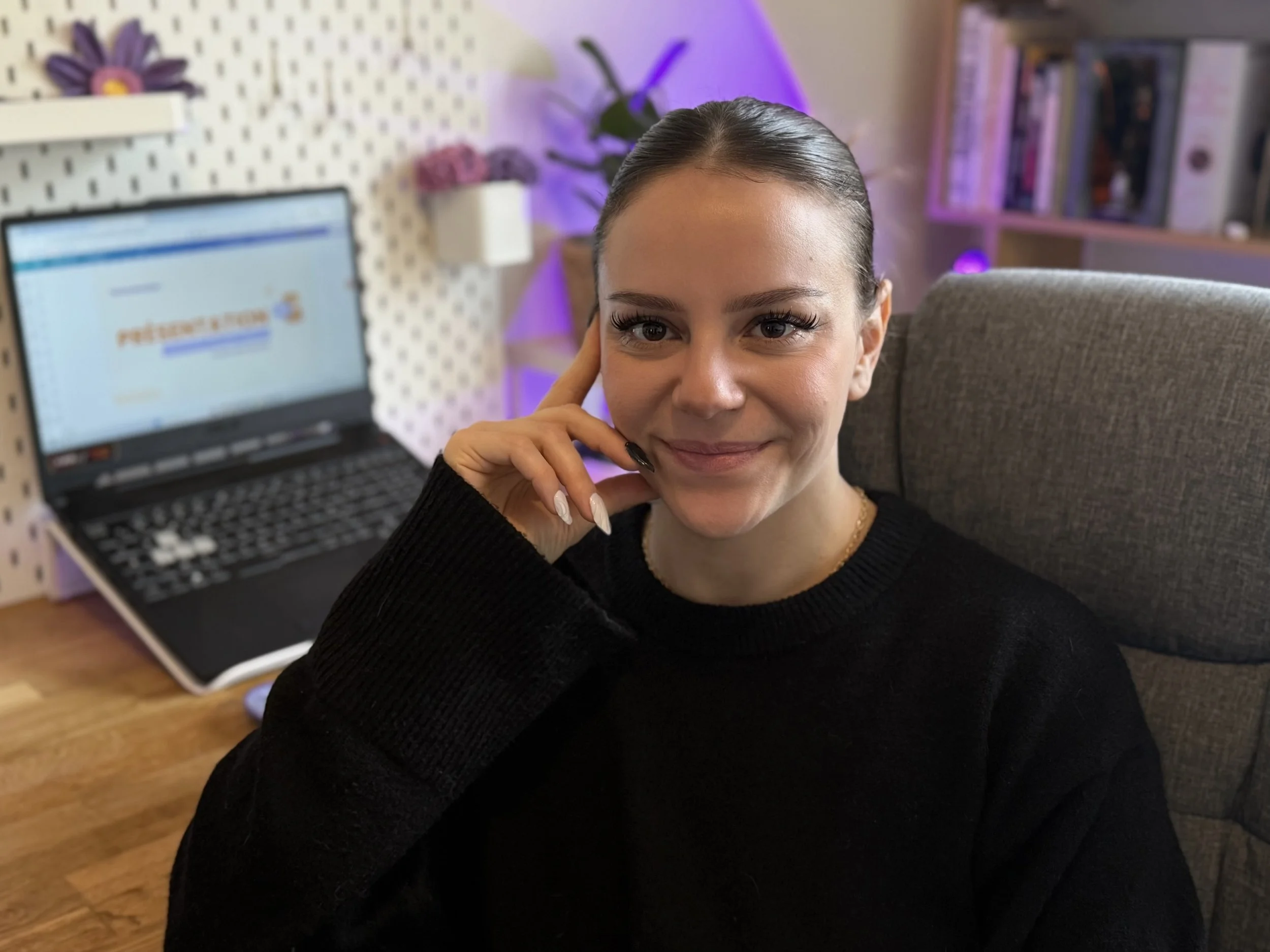 Une femme souriante assise dans un fauteuil, avec un ordinateur portable ouvert derrière elle, dans un bureau ou un espace de travail domestique, décoré de livres et de plantes.