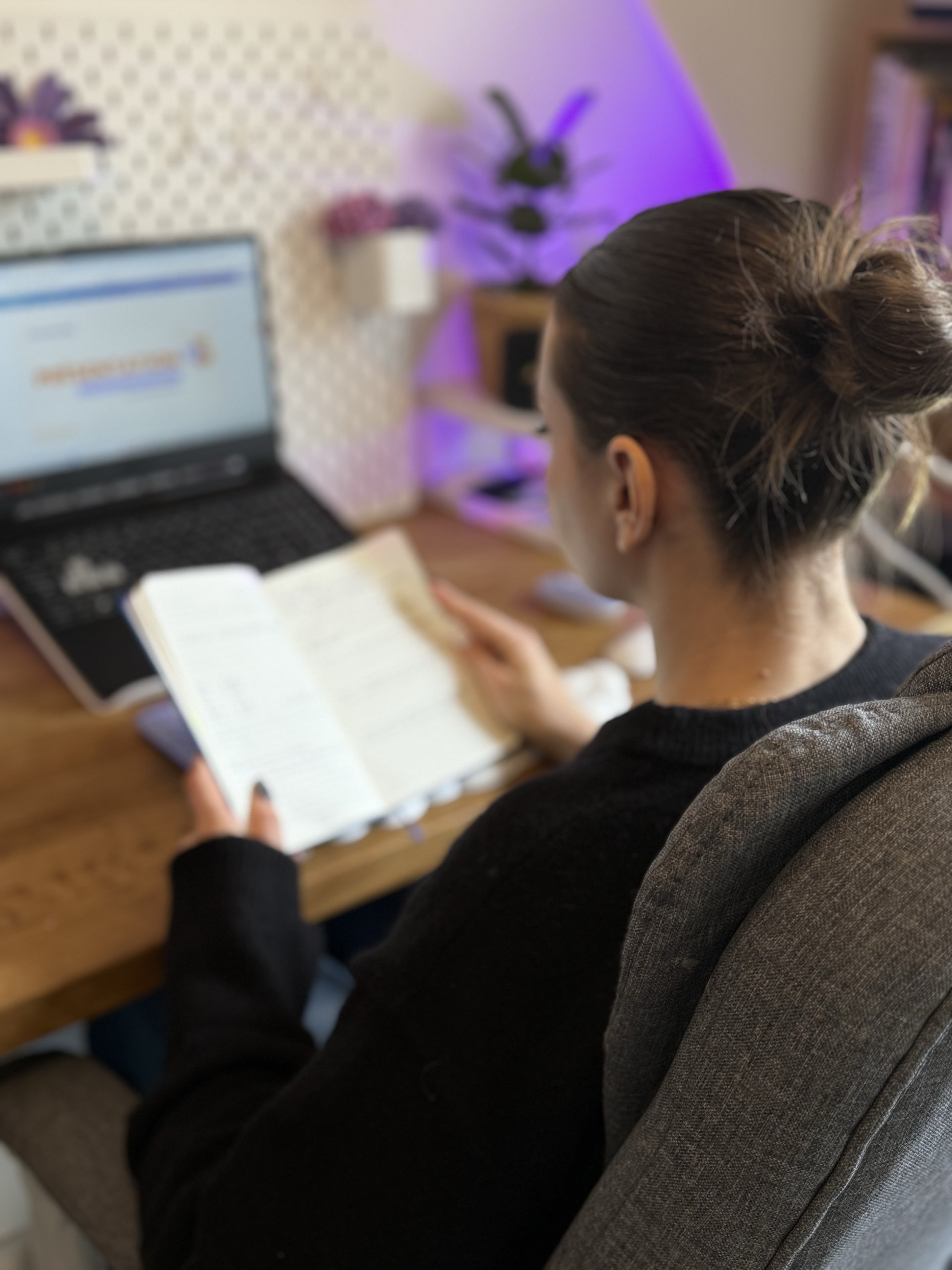 Une femme assise à un bureau regardant un livre, avec un ordinateur portable allumé à côté, dans un environnement avec des plantes et un éclairage violet