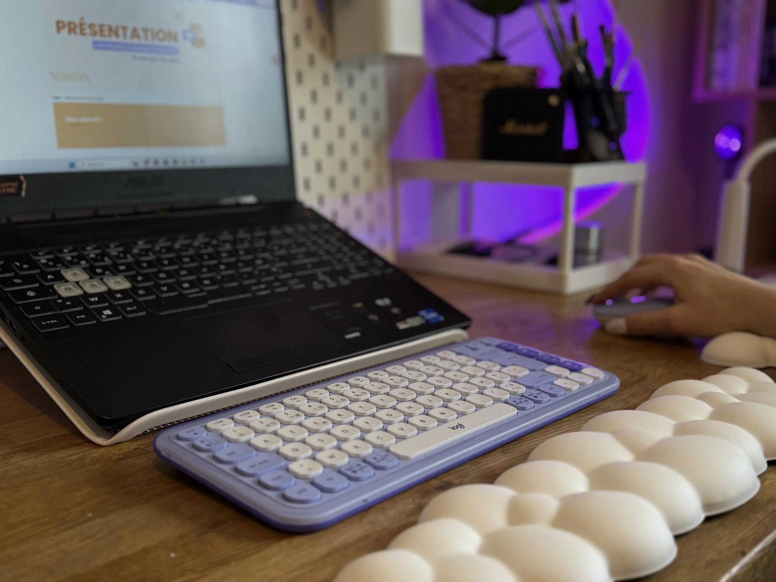 Espace de travail avec un ordinateur portable ouvert, un clavier compact, une souris, et une main utilisant la souris. En arrière-plan, une étagère avec des fournitures de bureau et un éclairage violet.