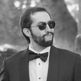 Man with sunglasses, beard, tuxedo, and bow tie outdoors
