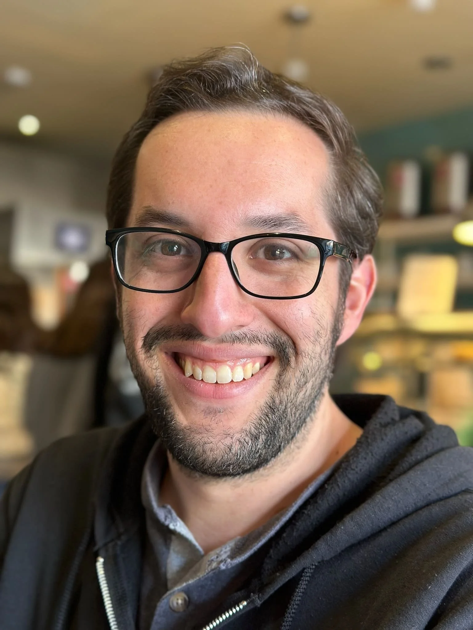 A smiling man with glasses and facial hair, wearing a dark hoodie, in a cozy indoor setting.