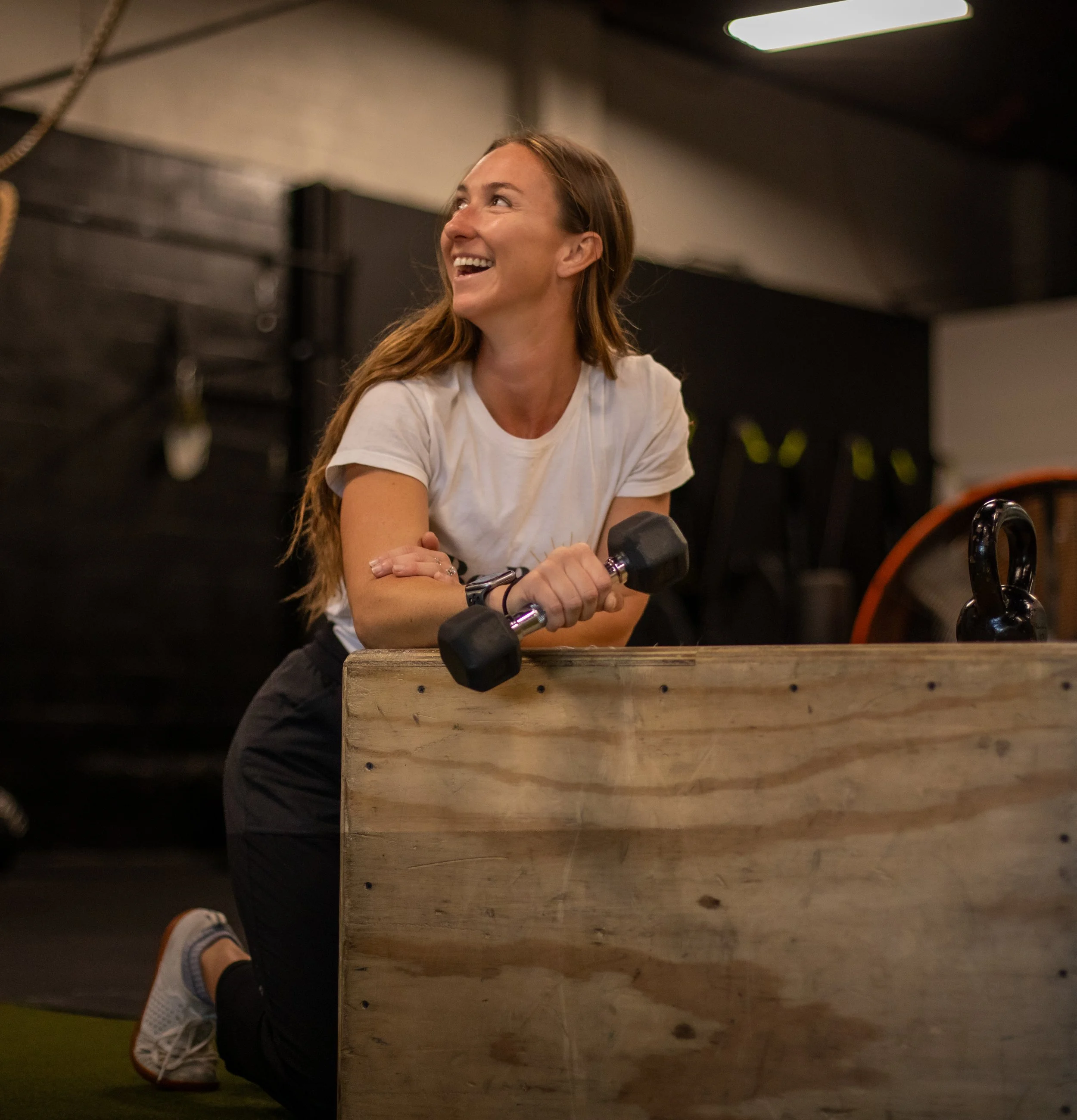A woman with long red hair, dressed in a white t-shirt and black workout pants, is at a gym, holding a dumbbell with one hand while leaning on a wooden box, smiling and looking to the side.