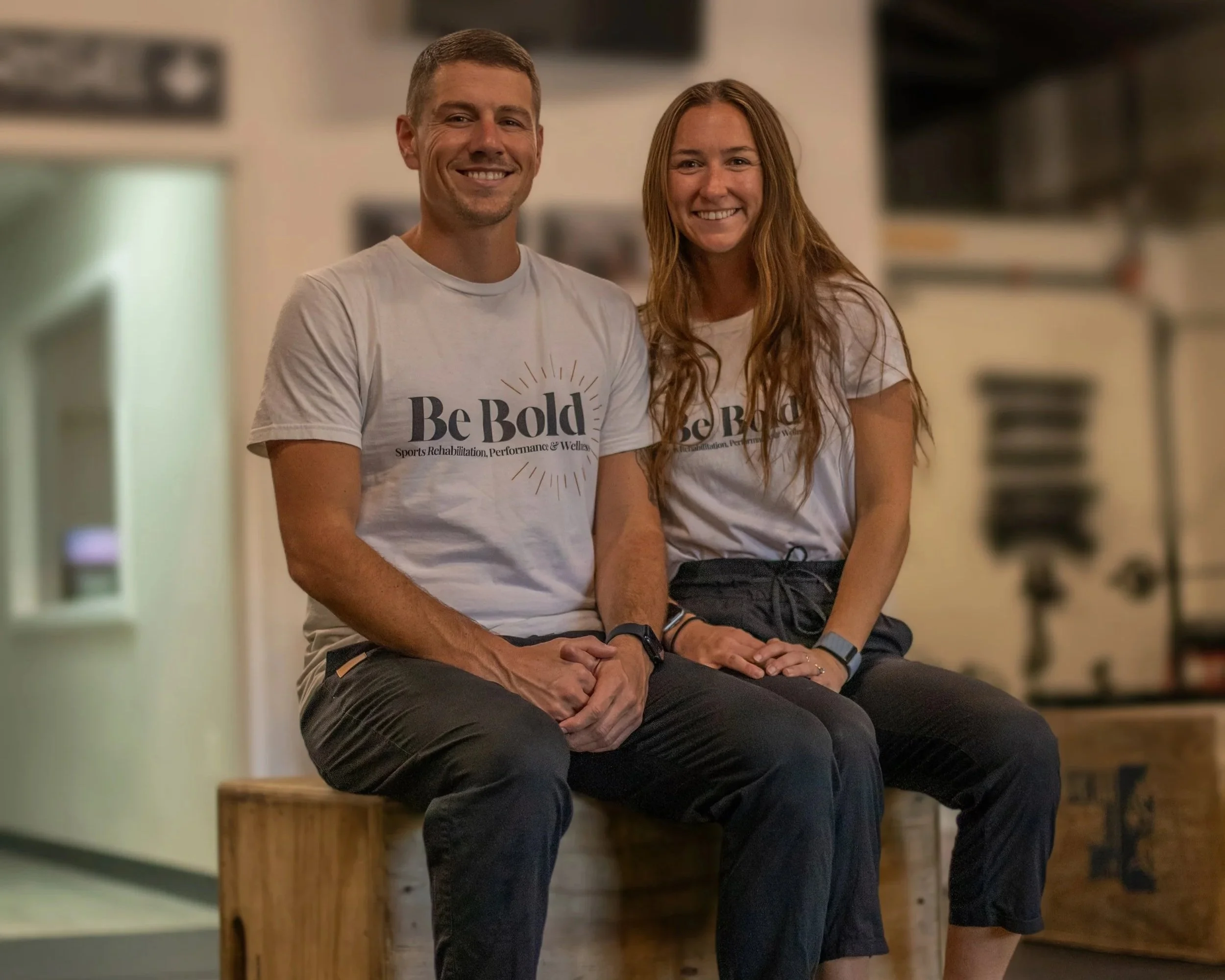 A smiling man and woman sitting on a wooden bench, both wearing white t-shirts with the text "Be Bold" and additional smaller text, in an indoor setting with blurred background.