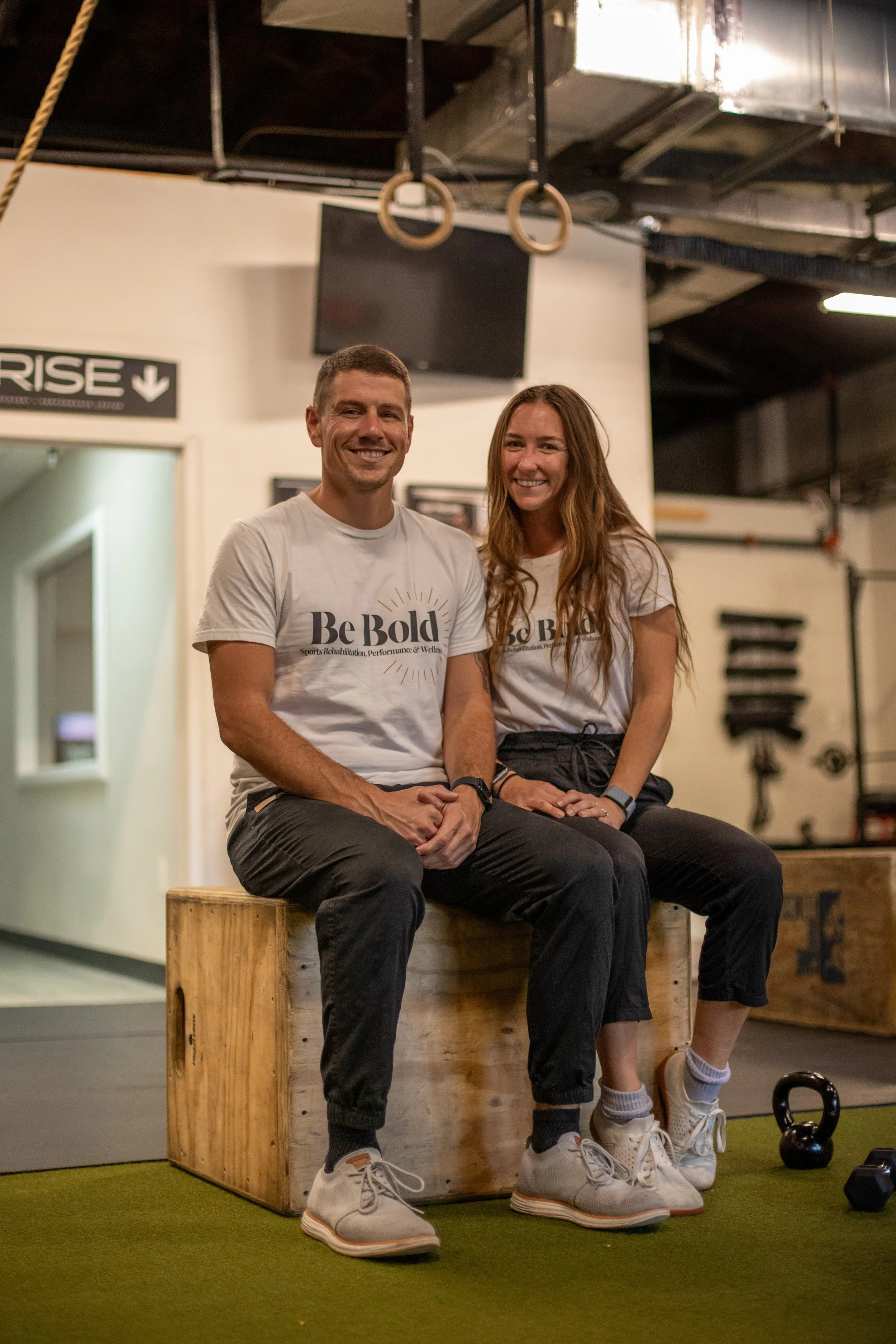 Two smiling people sitting on a wooden box inside a gym, wearing matching white t-shirts that say 'Be Bold.' There are gym equipment and rings hanging from the ceiling behind them.