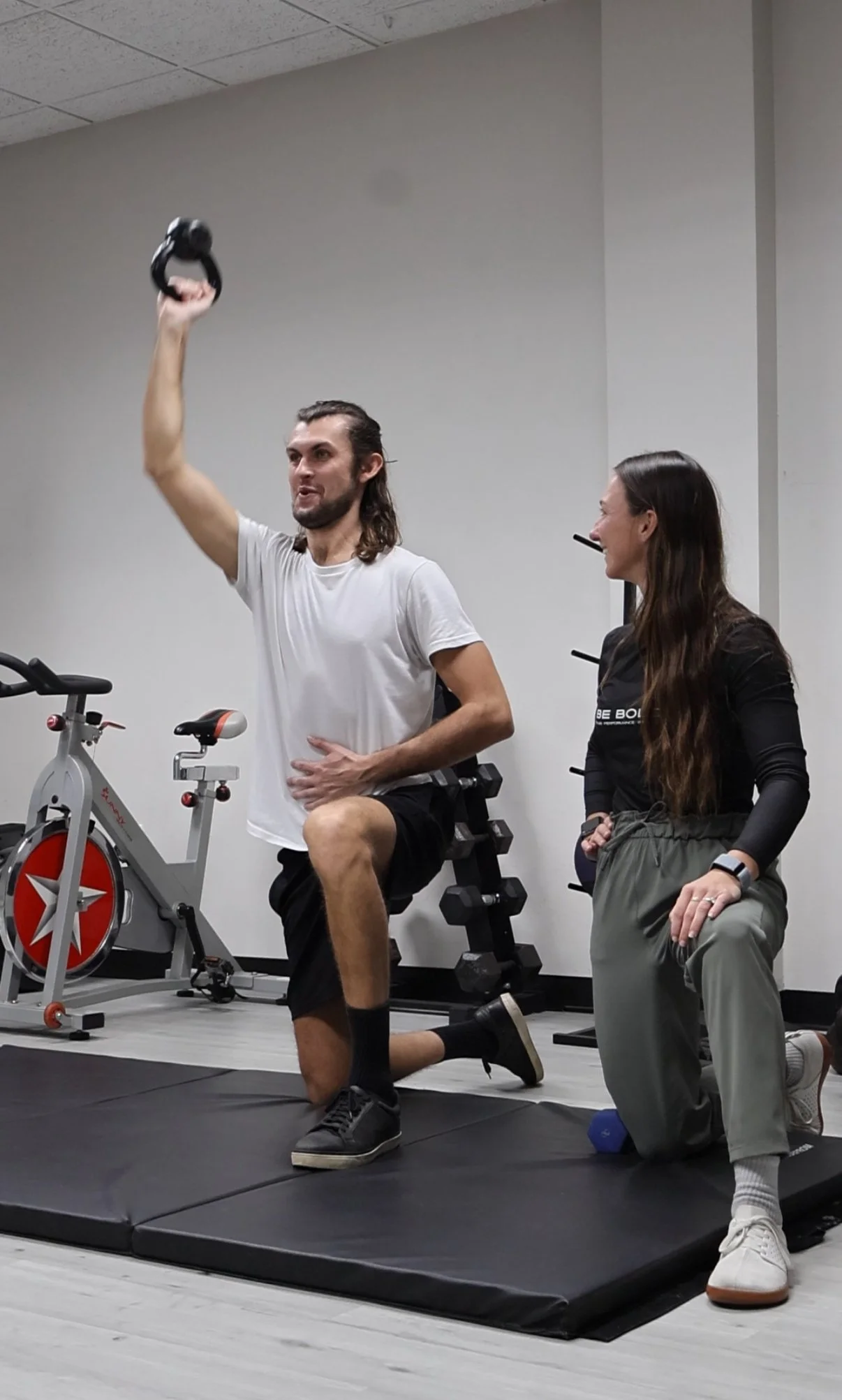 A man in a white t-shirt and black shorts performing a kneeling weightlifting exercise with a kettlebell in a fitness gym, assisted by a woman kneeling beside him.