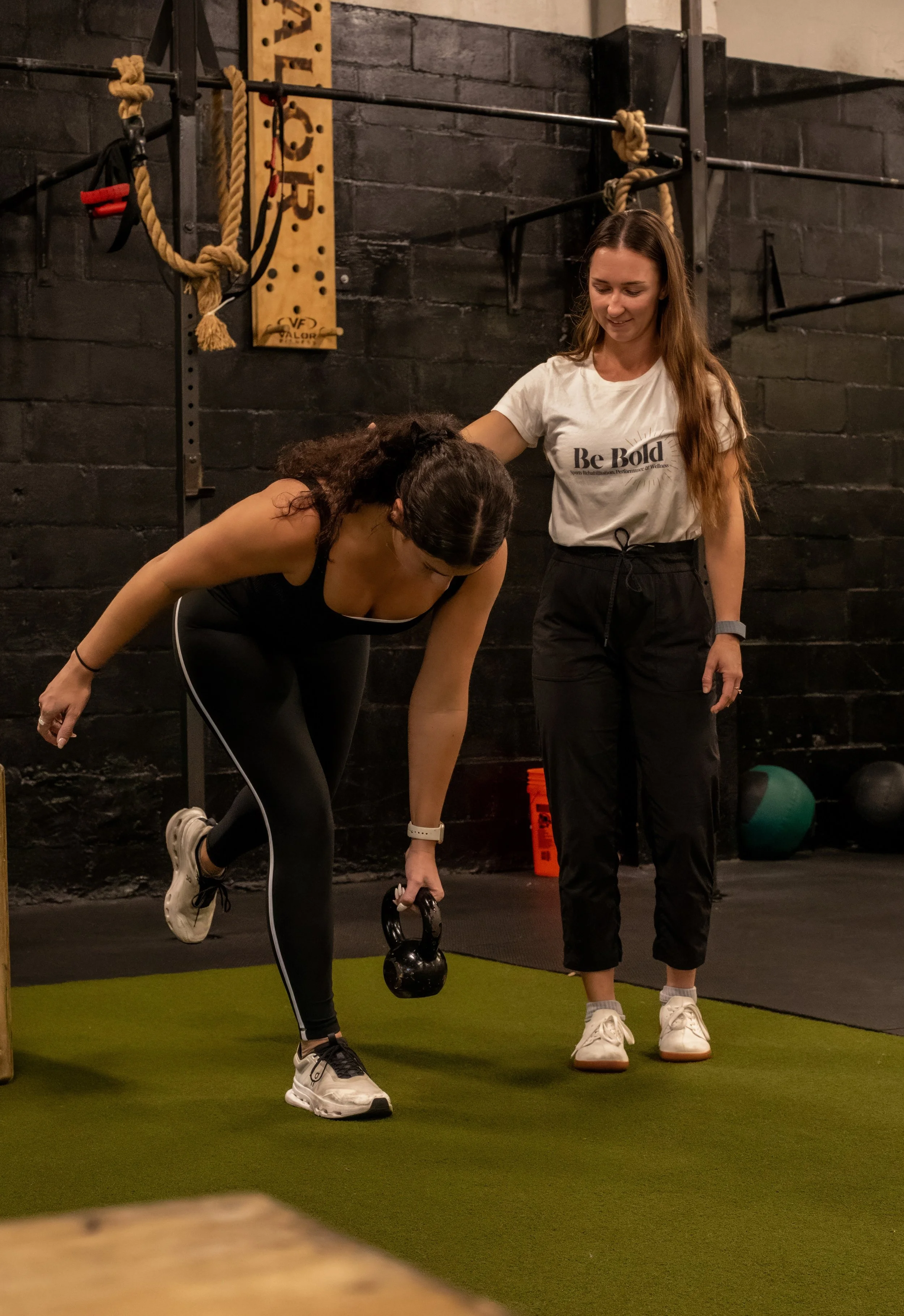 A woman performing a kettlebell exercise at a gym while a trainer guides her.