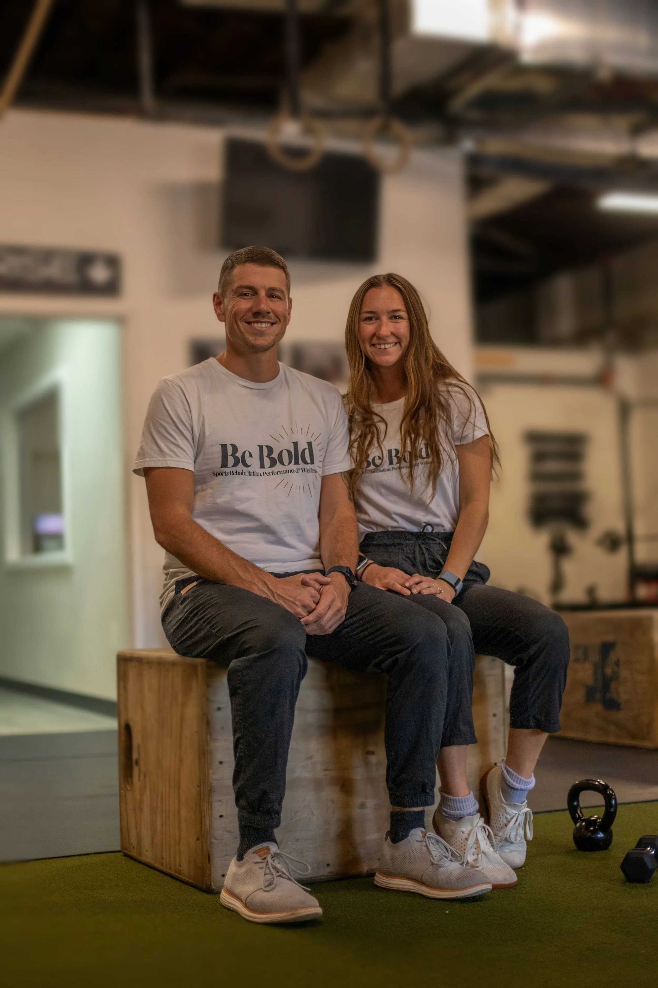Two smiling people, a man and a woman, sitting on a wooden box in a gym or fitness studio. They are wearing matching white T-shirts with the words 'Be Bold' printed on them, and black athletic pants. There are kettlebells and dumbbells on the floor nearby.