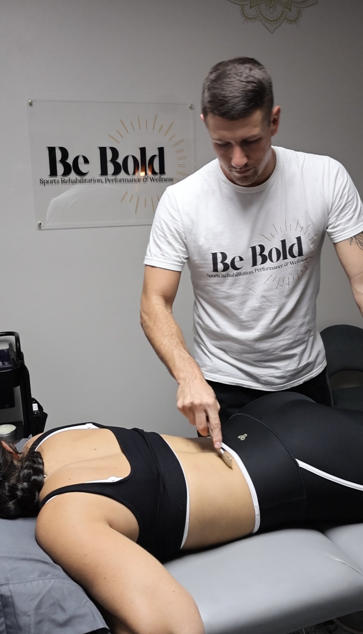 A man providing sports therapy or acupuncture to a woman lying face down on a treatment table. The man is wearing a white t-shirt with the words 'Be Bold' and a slogan about sports rehabilitation, performance, and wellness. The background has a similar logo on the wall.