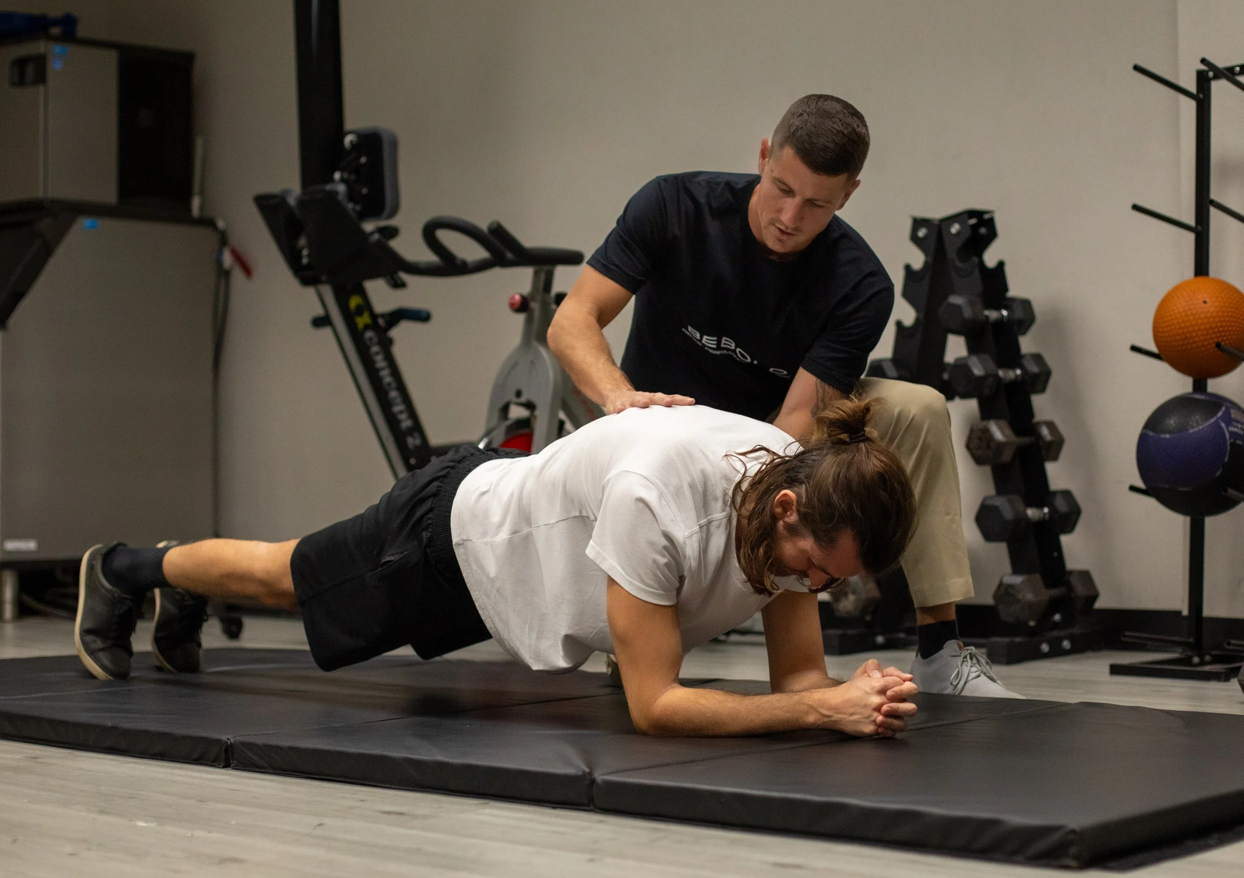 A man is instructing a woman doing a plank exercise on a padded mat in a gym, with fitness equipment in the background.