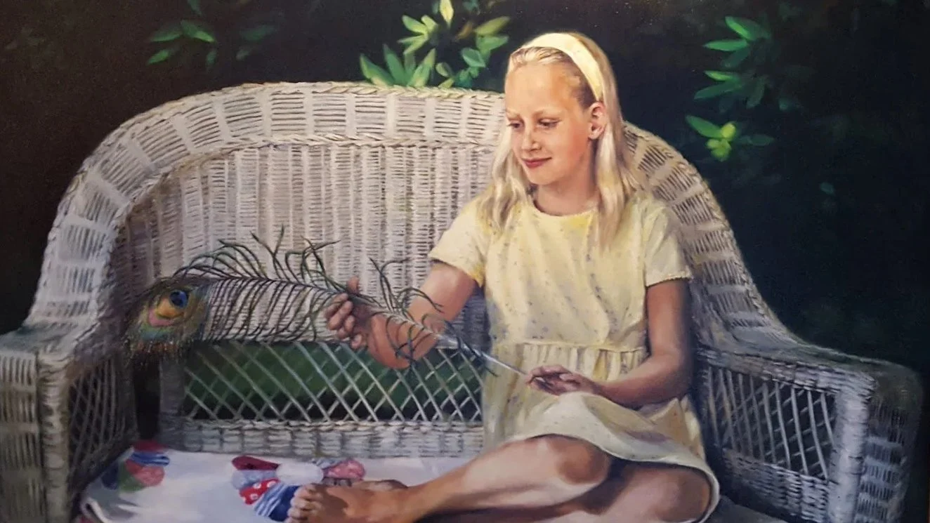 A young girl with long blonde hair sitting on a wicker bench holding a peacock feather, with a peacock feather design on her yellow dress, outdoors with greenery in the background.