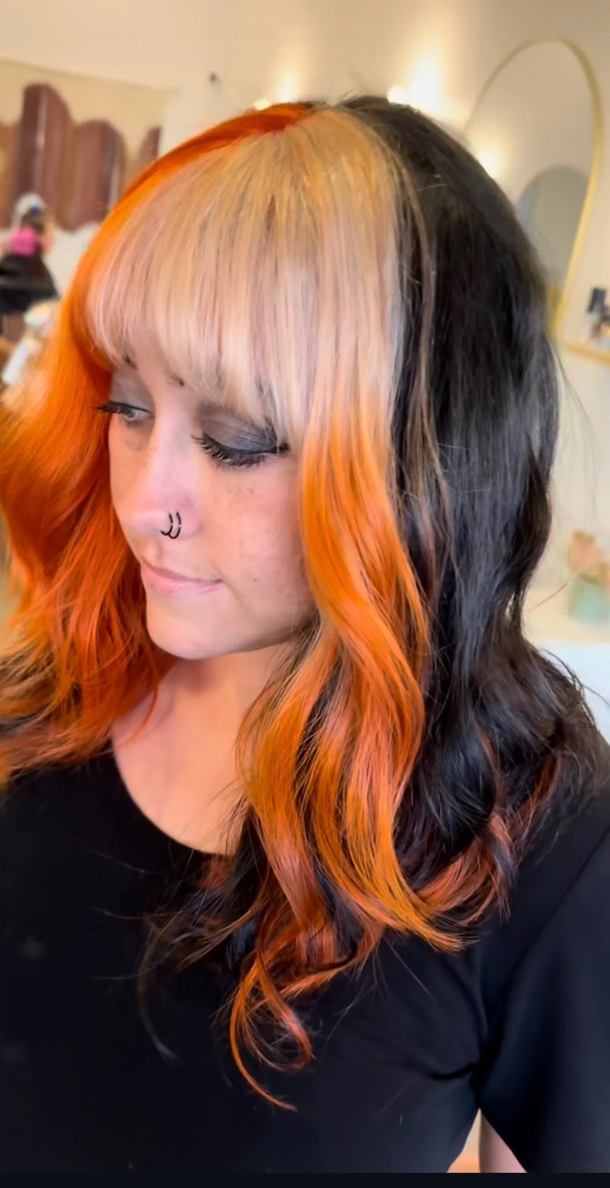  A woman with a bold split-color hairstyle featuring vivid orange-red on one side and jet black on the other, with a platinum blonde fringe bang, styled in soft waves in a modern hair salon.