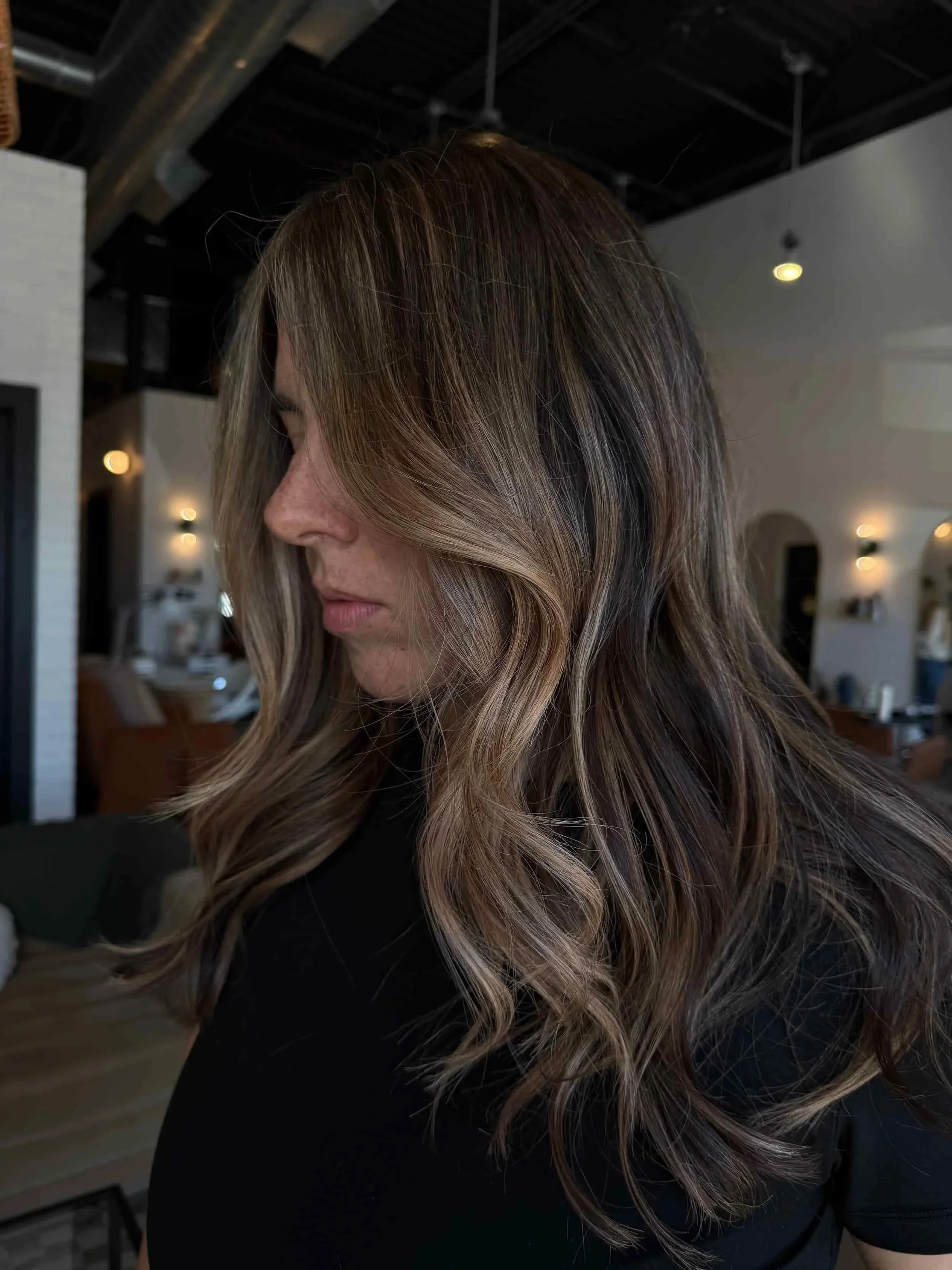 Back view of a woman with very long, rich dark brown hair featuring subtle bronze and caramel balayage highlights styled in defined beach waves, shown in a bright open hair salon.