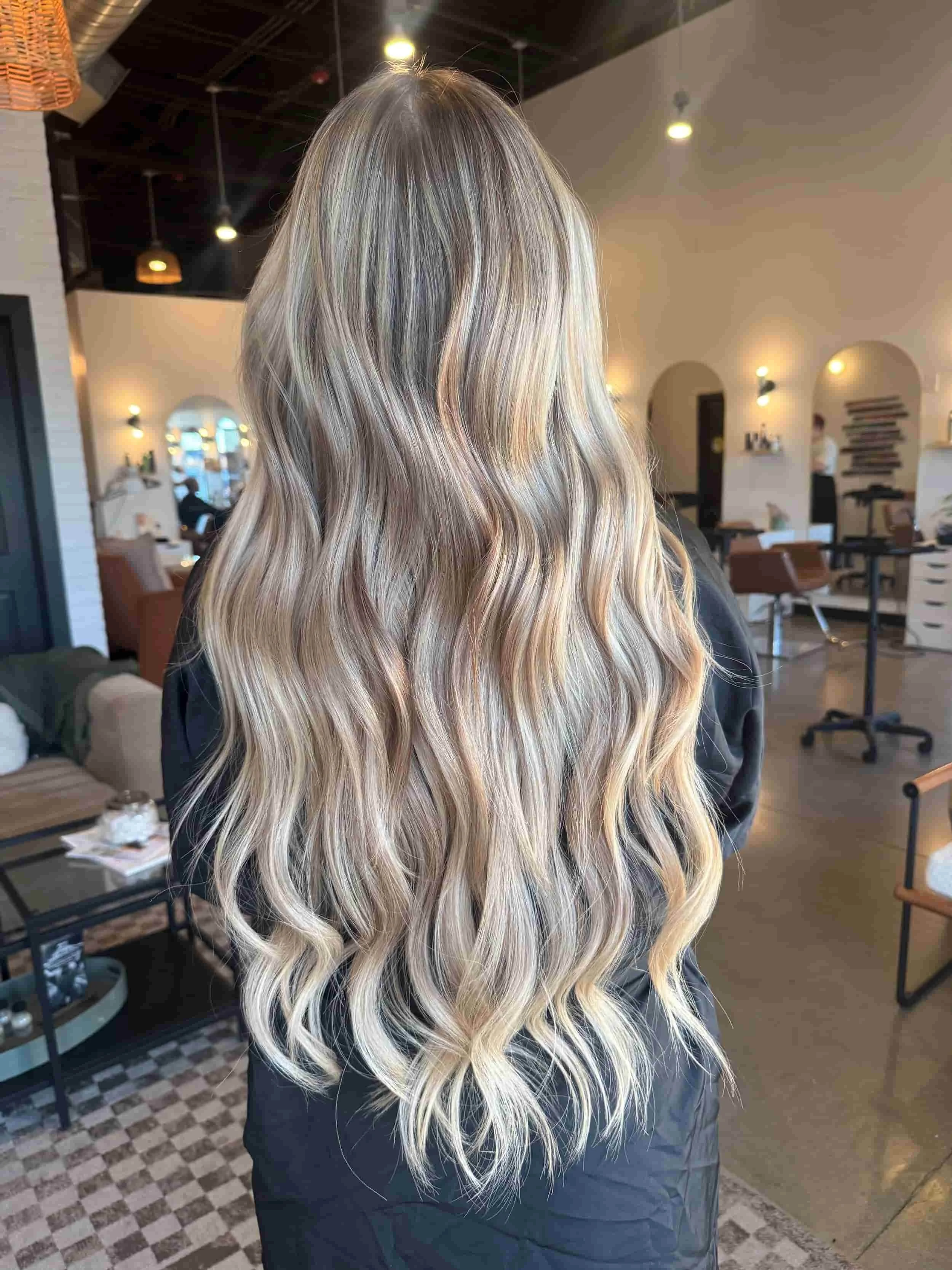 Back view of a woman with long, flowing sandy blonde hair featuring balayage highlights, a hybrid extension install and loose soft waves, styled in a modern salon with white brick walls and warm lighting.