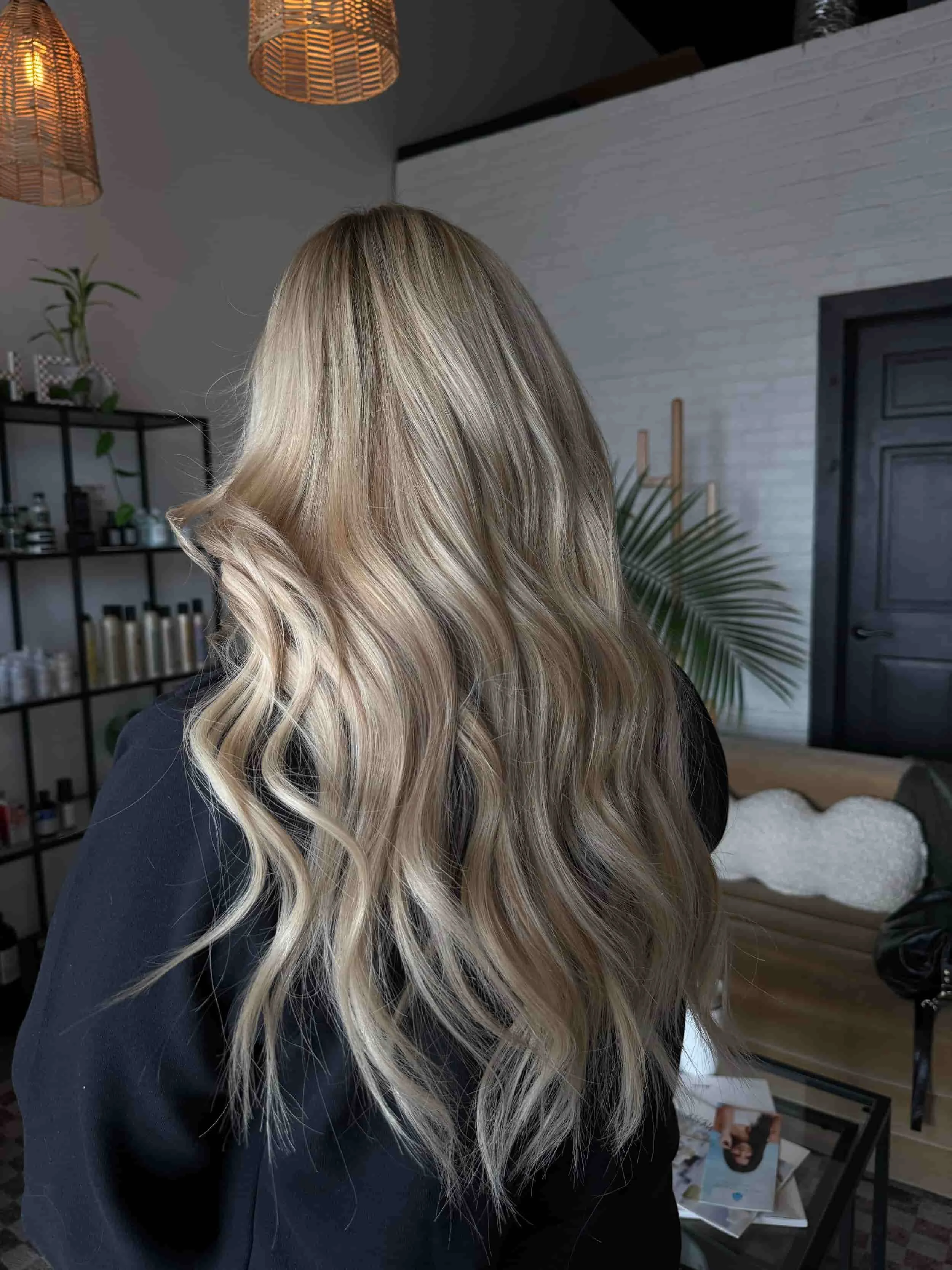 Back view of a woman with long, voluminous platinum blonde hair featuring dimensional highlights, k-tip extensions, and soft beach waves, photographed in a bright modern hair salon.