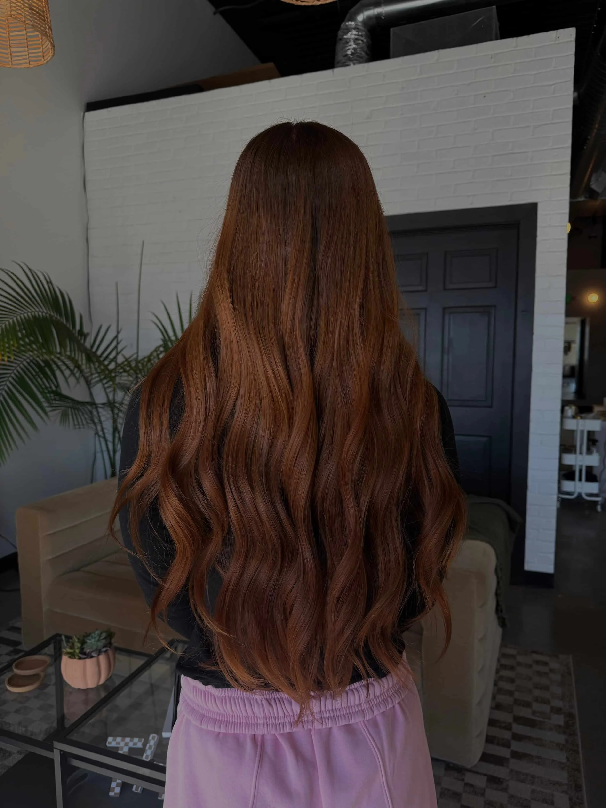 Back view of a woman with very long, rich auburn and cinnamon brown hair featuring subtle copper highlights and soft, flowing beach waves, photographed in a modern hair salon with white brick walls and warm lighting.